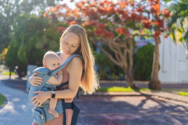 Bir anne, ergonomik bir bebek taşıyıcısı ile bebeğini vücuduna yakın bir şekilde dışarı çıkar. Bebek giyimi, aktif ebeveynlik, rahatlık, bağlılık, hareketlilik ve modern aile yaşam tarzı kavramı