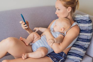Bebek akıllı telefonuna bakarken annesinin kucağında oturuyor, duygusal olarak kopuk. Perdenin bebekler ve karşılanmamış duygusal ihtiyaçlar üzerindeki etkisini gösteren sembolik bir sahne. Dijital ebeveynlik ve