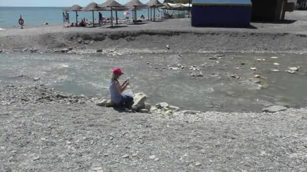 Sarışın kırmızı şapka ve güneş gözlüğü oturuyor bir sahil, dağ küçük dere üzerinde hiçbir Far From Pebble Beach giymiş — Stok video