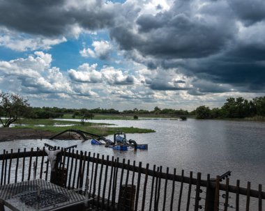 Geniş bir nehrin üzerinde, kısmen sular altında kalmış mavi bir traktör ve ön planda kırsal bir ahşap çitle yuvarlanan koyu renk fırtına bulutlarının dramatik görüntüsü. Sahne gerilimi ve kırsal kesimi çağrıştırıyor