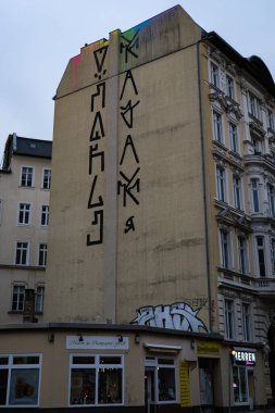 Yüksek dış duvarda kalın dikey graffitileri olan bir apartmanın sokak manzarası. Klasik Avrupa mimarisi ve küçük dükkanlarla çevrili sahne, otantik