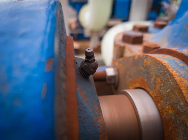 Close-up of a grease fitting (zerk) on rotating industrial machinery. Visible rust, bolts, and motion blur highlight active lubrication in a heavy-duty environment.