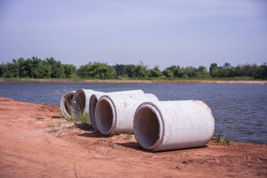 Beton drenaj boruları, büyük bir su deposunun yakınındaki kırmızımsı topraklara döşenmiş. İnşaat, altyapı, inşaat mühendisliği ve çevre planlaması için ideal.