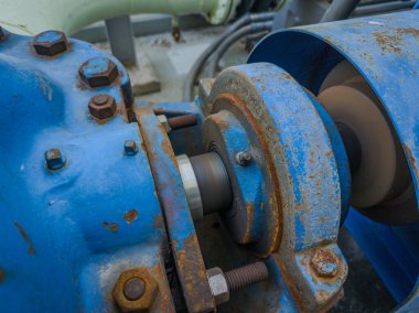 Close-up of a grease fitting (zerk) on rotating industrial machinery. Visible rust, bolts, and motion blur highlight active lubrication in a heavy-duty environment