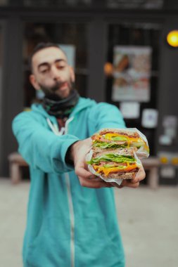 Genç sakallı yetişkin adam restoranın önünde büyük porsiyon sandviç tutuyor. Sağlıklı gıda konsepti, yakın plan, ön görüş.