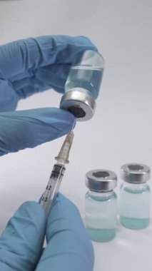 Mavi eldiven giyen bir sağlık uzmanı aşı şişesinden şırıngayla sıvı alır. Arkaplan şişelerinde hazırlık ve güvenlik vurgulandı.