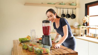 Smiling woman making a colorful fruit smoothie in a blender at a bright kitchen setting on the counter, surrounded by fruit and vegetable.