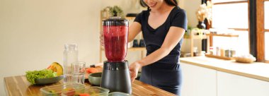 Smiling woman making a colorful fruit smoothie in a blender at a bright kitchen setting on the counter, surrounded by fruit and vegetable.