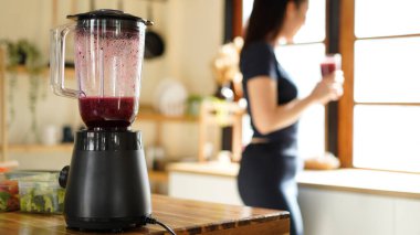Black electric blender making a smoothie with raspberries, blueberry, with a woman preparing healthy food in the background.
