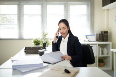 businesswoman talking on smartphone and reading document, solving corporate business problem. Female employee discussing document by cellphone, receive news..