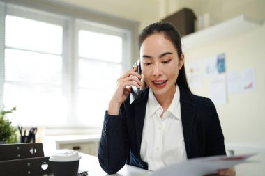 businesswoman talking on smartphone and reading document, solving corporate business problem. Female employee discussing document by cellphone, receive news..