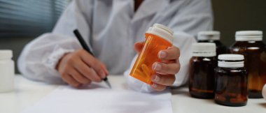 Woman doctor reviews medication bottle while writing notes in a sunlit clinic room suggesting a professional medical workplace setting..