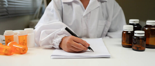 Close-up of a doctor or pharmacist's hands writing a prescription on a clipboard with medicine bottles in the background.