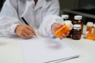 Woman doctor reviews medication bottle while writing notes in a sunlit clinic room suggesting a professional medical workplace setting..