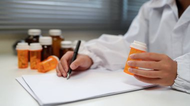 Woman doctor reviews medication bottle while writing notes in a sunlit clinic room suggesting a professional medical workplace setting..