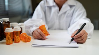 Woman doctor reviews medication bottle while writing notes in a sunlit clinic room suggesting a professional medical workplace setting..