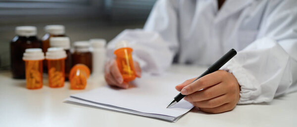Woman doctor reviews medication bottle while writing notes in a sunlit clinic room suggesting a professional medical workplace setting..
