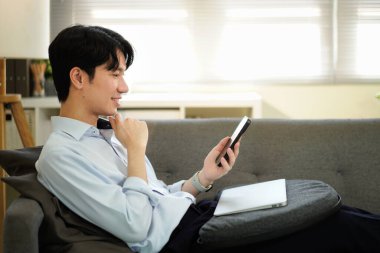 Sosyal ağ kurma, akıllı telefonu koltukta uzanan ve evinde oturma odasında yatan adam. Bağlantı ya da teknoloji, gülümseme ve cep telefonuyla online oyun oynayan mutlu bir erkek..