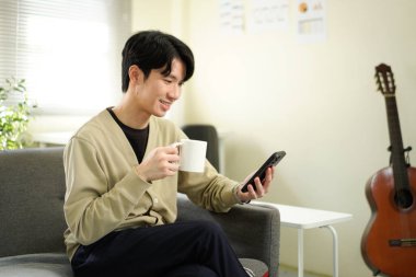 Rahat adam kanepede kahve, telefon, rahat ofis titreşimleri. Doğal Işıklı Sıradan Oturma Odası Sahnesi.