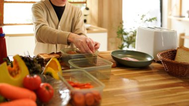 Bir adam mutfakta sandviç hazırlıyor. Ekmeği kesmek için bıçak kullanıyor ve sandviçleri plastik kaplara koyuyor..
