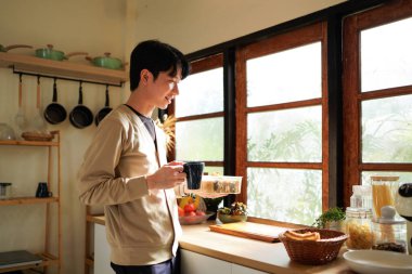 Bir adam sabah pencerenin yanında sandviç yiyor ve kahve içiyor. Sahne rahat ve sıradan..