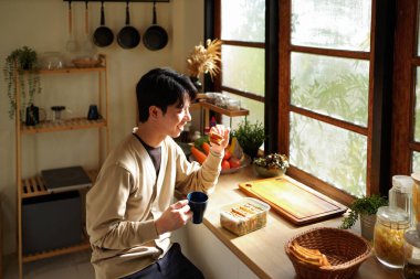 Bir adam sabah pencerenin yanında sandviç yiyor ve kahve içiyor. Sahne rahat ve sıradan..