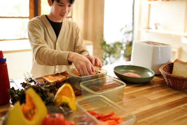 Bir adam mutfakta sandviç hazırlıyor. Ekmeği kesmek için bıçak kullanıyor ve sandviçleri plastik kaplara koyuyor..