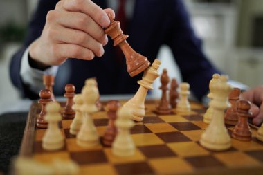 Satranç tahtasının üzerinde tahta satranç taşı tutan kişi kıyafeti diğer sarı satranç taşlarıyla odaklanmış, stratejik düşünce ve konsantrasyonu gösteriyor..