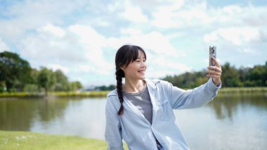 Bir kadın parkta dikilirken cep telefonuyla selfie çekiyor. Park ördekler ve diğer kuşlarla dolu ve kadın fotoğrafı çekerken gülümsüyor..