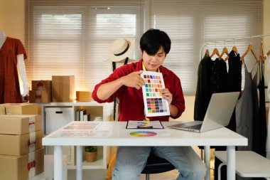 Genç tasarımcının portresi şık oturma ve çalışma tarzı. Genç adam moda tasarım stüdyosunda dizüstü bilgisayar ve renkli kumaşlar kullanıyor..