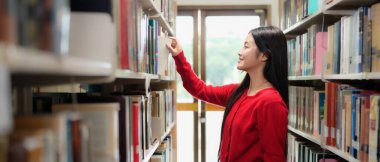 Üniversitede kütüphanede kitaplıkta kitap arayan öğrencilerin yan görünümü.