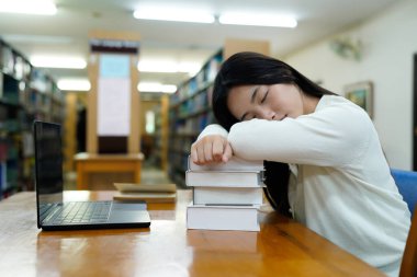 Kütüphanedeki bir masada uyuyan yorgun bir öğrenci. Yanında dizüstü bilgisayar ve kitaplarla...