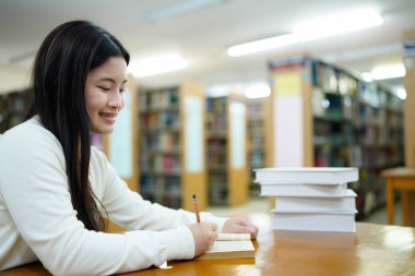 Üniversite ya da üniversite kütüphanesinde ödevlerini yapan Asyalı genç öğrenci kitaplığın arka planında çeşitli kitap ve sabit kitaplarla okula geri dönüyor..