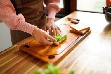 Kadın evde sandviç hazırlıyor. Sıcak, doğal bir mutfakta jambon ve taze sebzeler katlıyor..