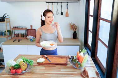 Mutlu Asyalı fitness kadını kulaklıkla müzik dinliyor ve mutfakta sebze salatası hazırlıyor, sağlıklı bir yaşam tarzının keyfini çıkarıyor..
