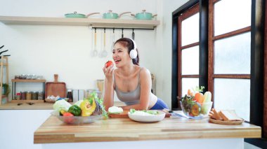 Mutlu Asyalı fitness kadını kulaklıkla müzik dinliyor ve mutfakta sebze salatası hazırlıyor, sağlıklı bir yaşam tarzının keyfini çıkarıyor..