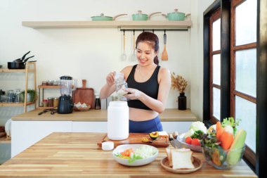 Kilo kaybı için diyet yemeği, spor giyimli Asyalı genç kadın, el ele tutuşup mutfakta protein şişesi yapıyor..
