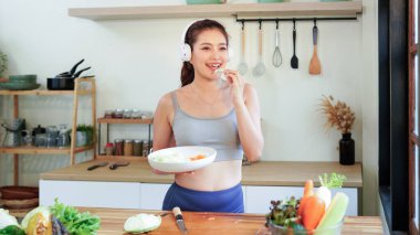 Mutlu Asyalı fitness kadını kulaklıkla müzik dinliyor ve mutfakta sebze salatası hazırlıyor, sağlıklı bir yaşam tarzının keyfini çıkarıyor..