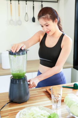 Mutfakta yeşil smoothie 'yi karıştıran, spor kıyafetleri giyen, tezgahta taze meyvelerle sağlıklı bir içecek hazırlayan, sağlıklı bir yaşam tarzına odaklanan bir kadın..