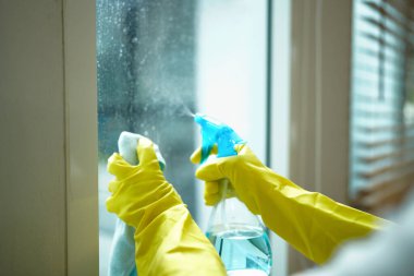 Sarı eldivenli biri pencereyi sprey ve bezle temizliyor..