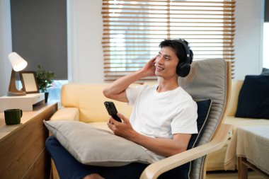 Kulaklık takmış gülümseyen genç adam akıllı telefon uygulamasında cep telefonu müziği dinliyor, mutlu adam telefonu tutarak rahatlıyor ve favori şarkıların tadını çıkarıyor..