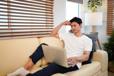 Genç bir adam oturma odasındaki kanepede dizüstü bilgisayar kullanıyor..