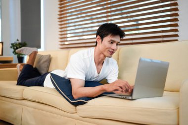 Modern evdeki rahat kanepeye uzanmış dizüstü bilgisayar kullanan kendine güvenen Asyalı adam..