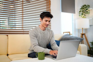 Genç Asyalı erkek kullanıcı dizüstü bilgisayar tutarak rahatlıyor ve oturma odasında ekrana bakıyor, uzaktan kumandalı iş ya da ev konseptinden çalışma..