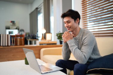 Genç Asyalı erkek kullanıcı dizüstü bilgisayar tutarak rahatlıyor ve oturma odasında ekrana bakıyor, uzaktan kumandalı iş ya da ev konseptinden çalışma..