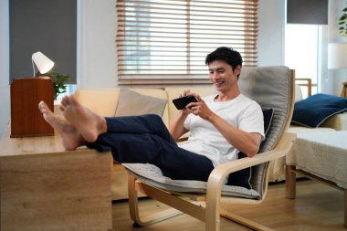Rahat genç Asyalı bir adam oturma odasında rahat bir şekilde otururken akıllı telefon kullanıyor..