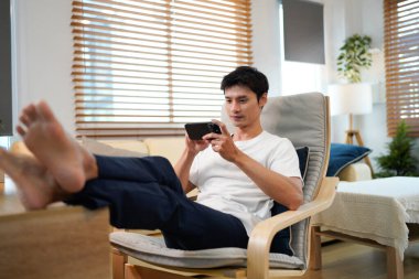 Rahat genç Asyalı bir adam oturma odasında rahat bir şekilde otururken akıllı telefon kullanıyor..