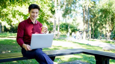 Parkta dizüstü bilgisayarda çalışan, parktaki bankta kahve içen genç bir adam..