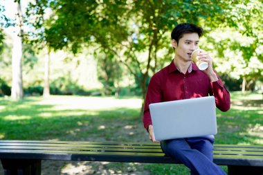 Parkta dizüstü bilgisayarda çalışan, parktaki bankta kahve içen genç bir adam..