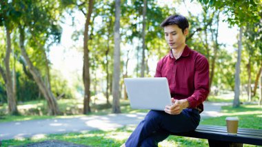 Parktaki bankta oturan bir adam yanında kahve fincanıyla dizüstü bilgisayar kullanarak güneşli bir günün tadını çıkarıyor..